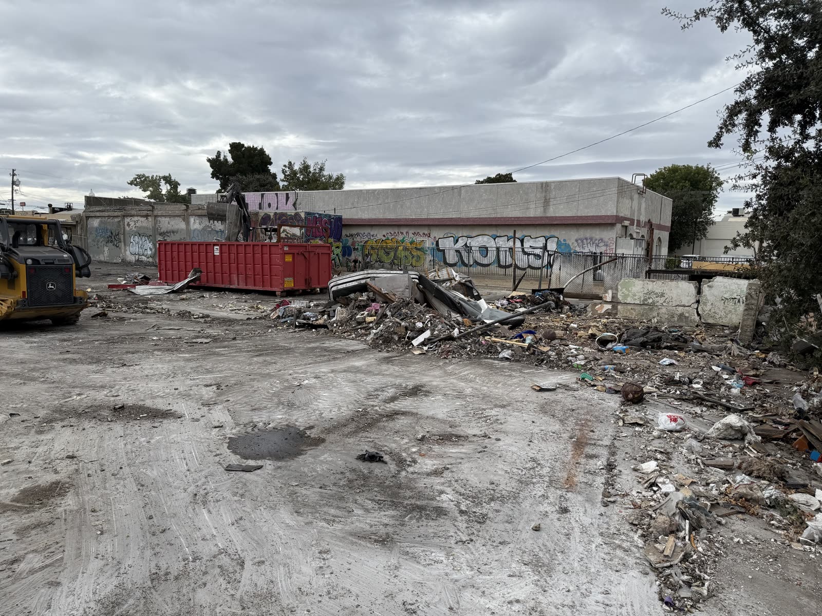 Demolition debris sorted across the slab beside a roll-off bin, with the remaining tagged wall behind
