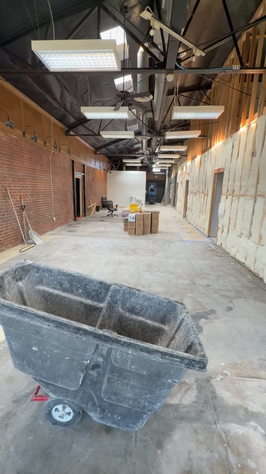 Walkthrough of an open commercial shell with exposed brick walls, an open ceiling with new ductwork and lighting, and floor protection down
