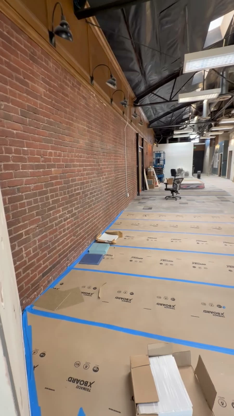 Commercial interior mid build-out with exposed brick, temporary lighting and taped floor protection covering the finished slab