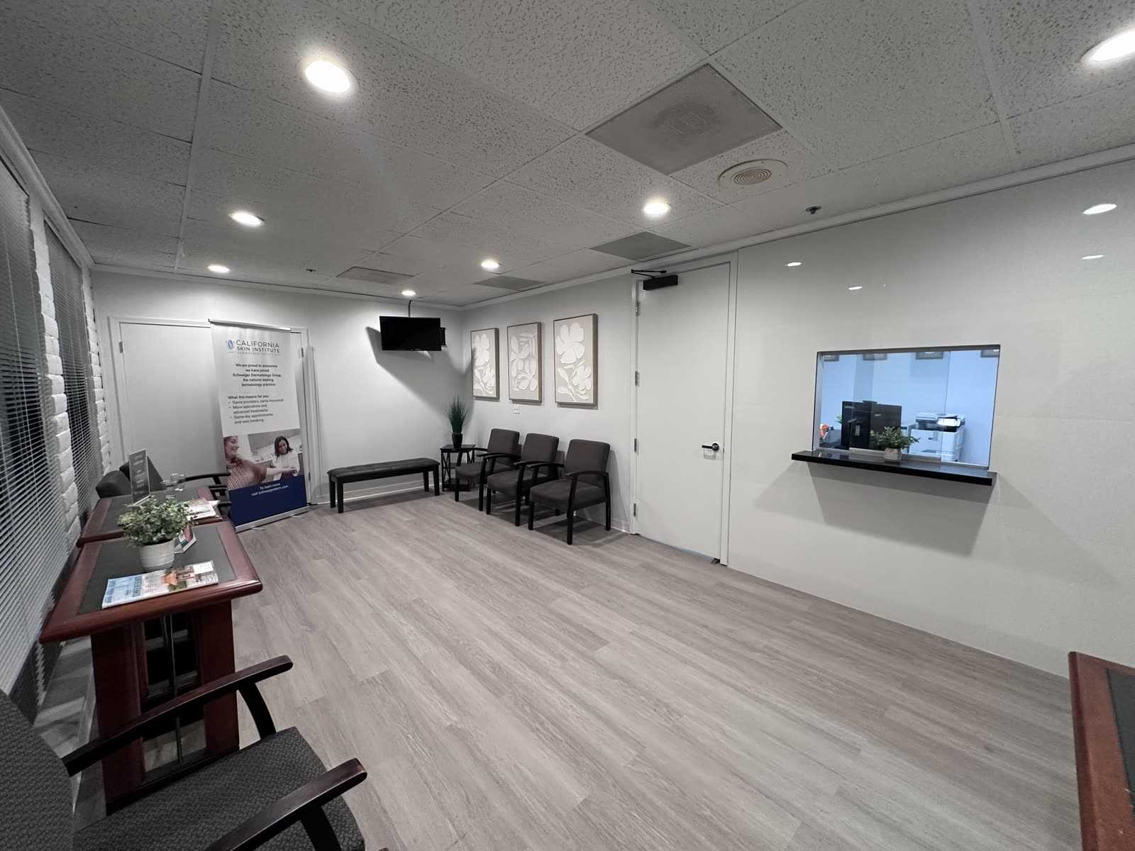 Dermatology clinic reception area with a glazed reception window, wall-mounted screen and framed artwork above patient seating
