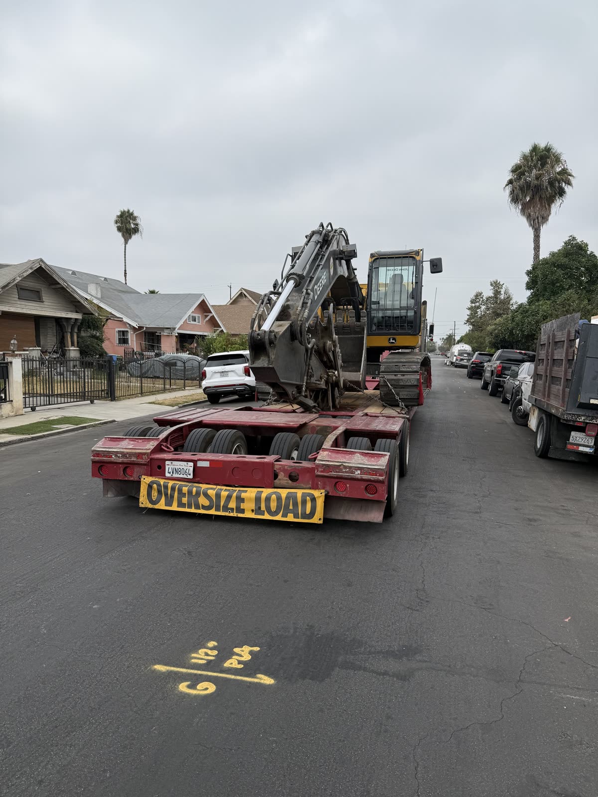 Excavator loaded on a lowboy trailer with an oversize load banner, staged in the street outside a residential demolition job