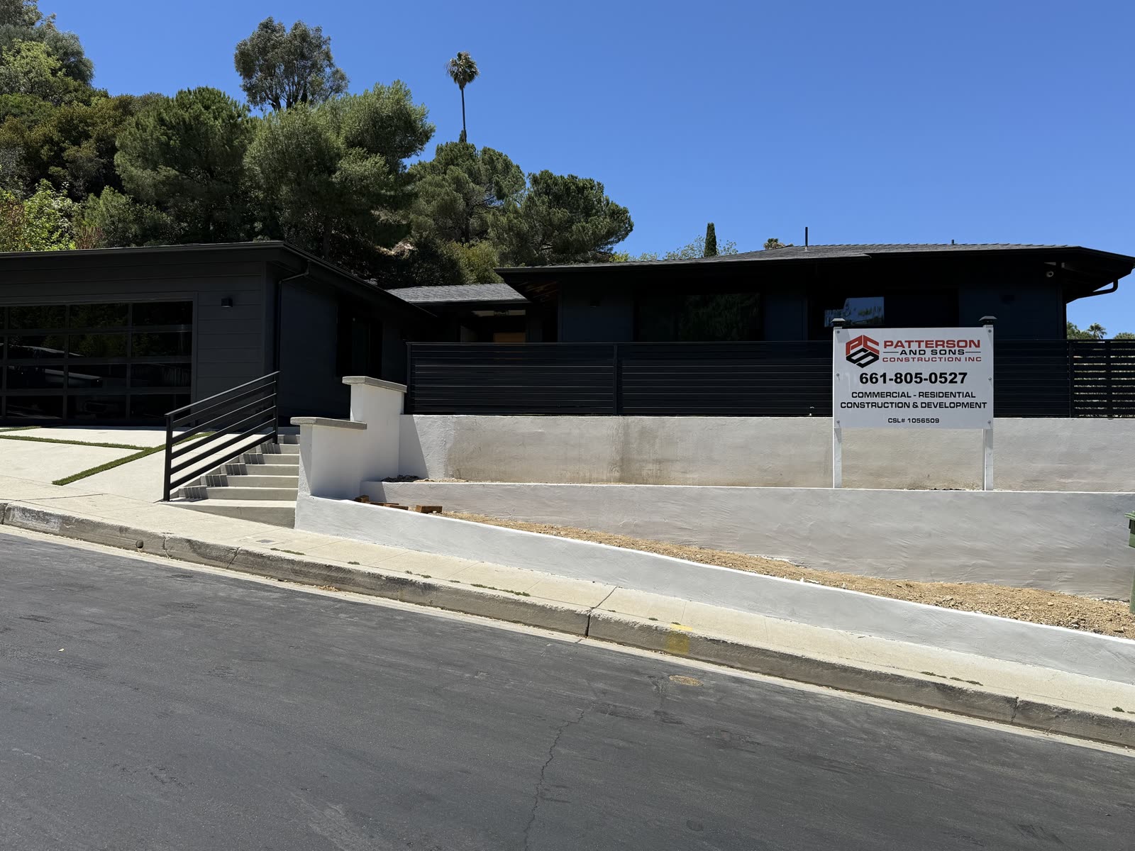 Renovated hillside home from the street with new board-formed stucco site walls, concrete steps, steel handrail and a horizontal slat privacy fence