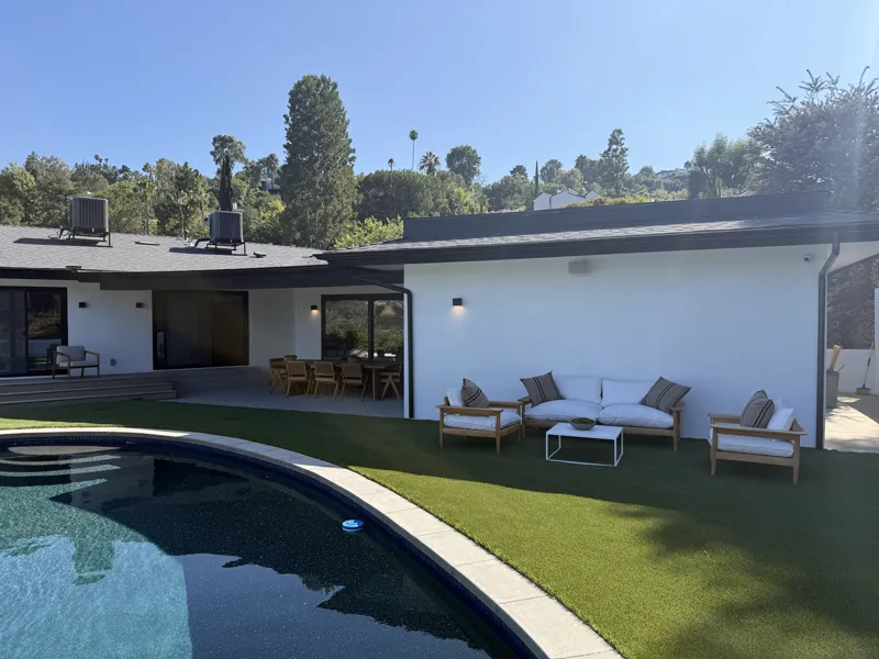 Renovated hillside home with white stucco, black window frames, a covered patio and artificial turf running to the pool coping