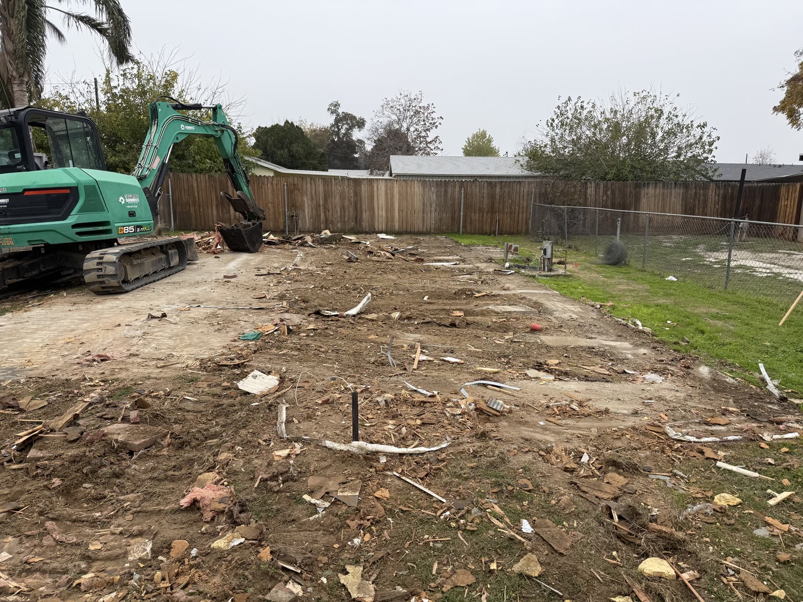 Compact excavator clearing the last debris off the pad after a mobile home was removed from a back yard