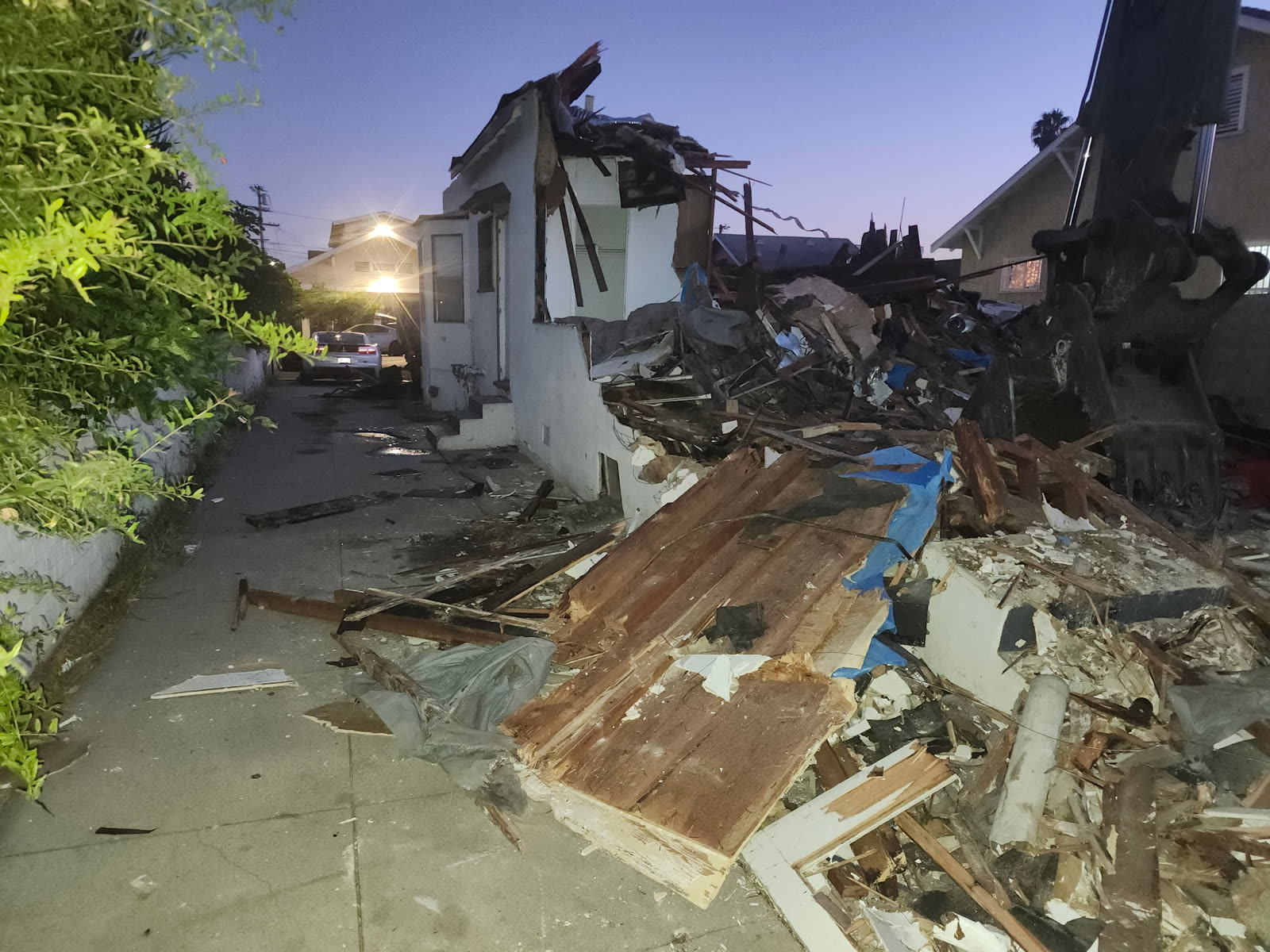 Partially demolished house at dusk with the rear half down, the excavator arm in frame and the side yard kept clear for access
