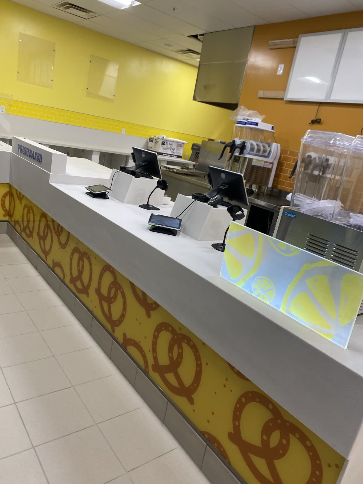 Completed food-court service counter with a quartz top, printed pretzel graphic panel, point-of-sale terminals and a frozen drink machine
