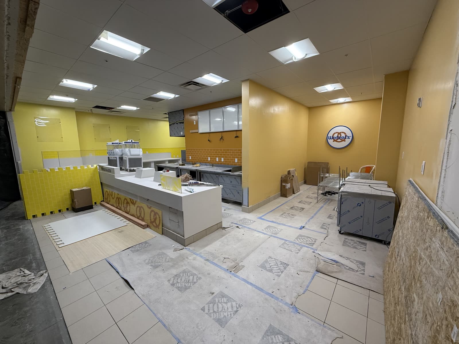 Food-court pretzel storefront near completion with the service counter set, wall tile going in and floor protection still down